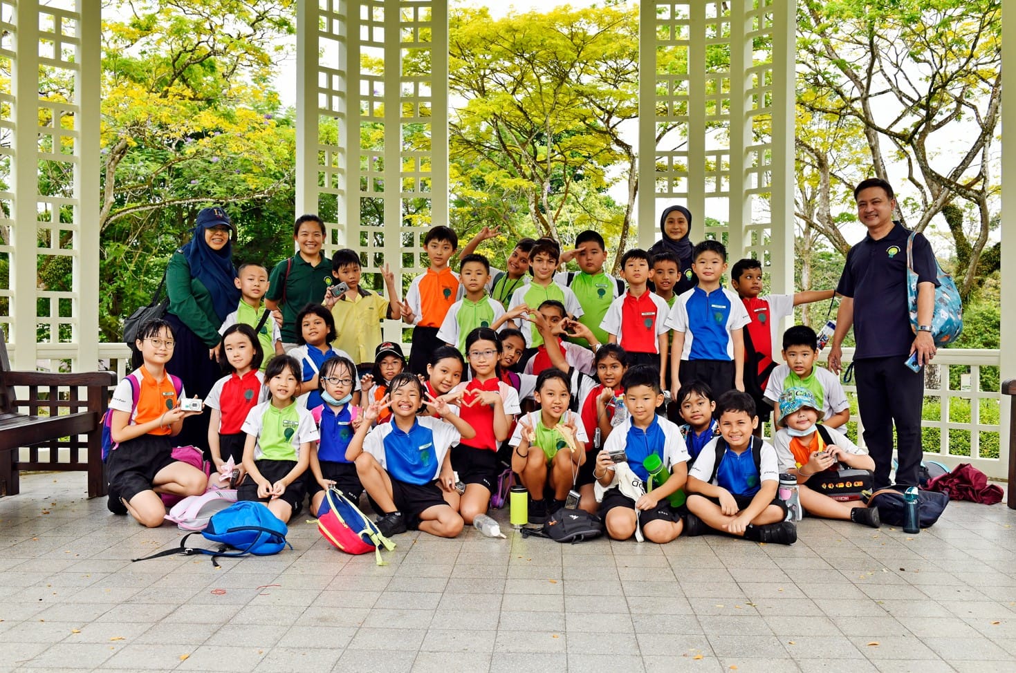A large group of young students and teachers posing together under an open white lattice gazebo surrounded by trees.